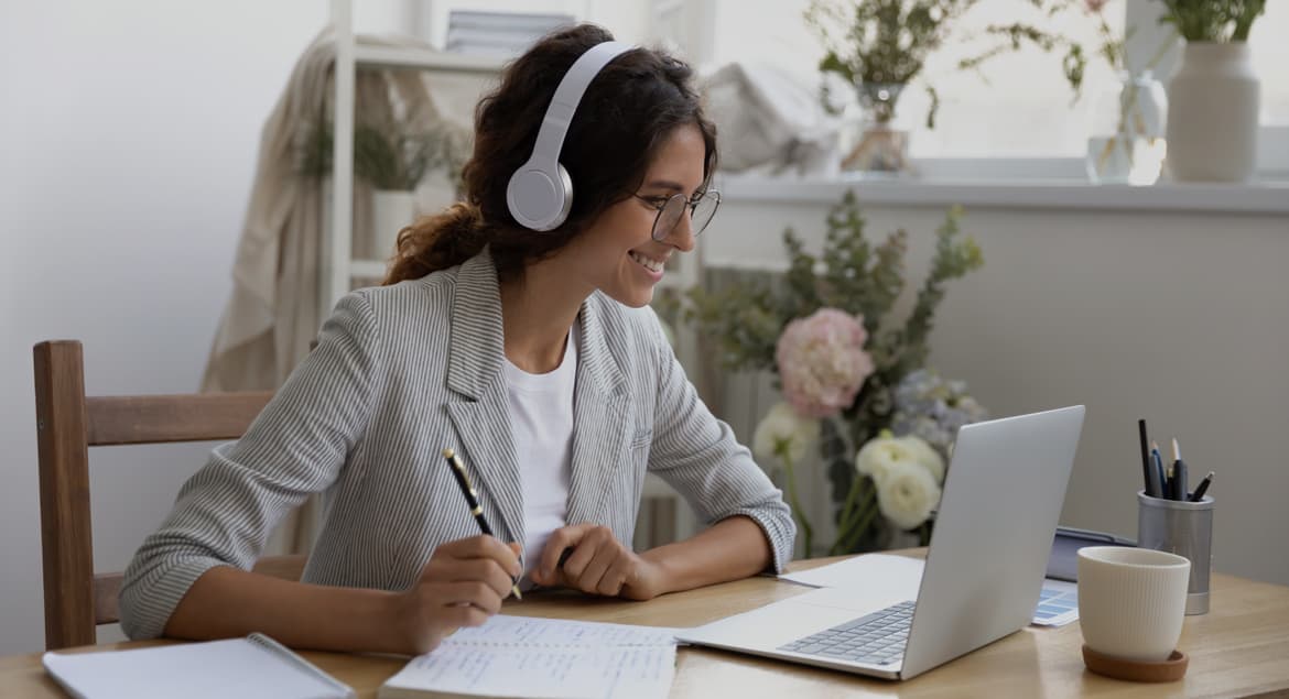 A woman participating in a virtual meeting with a laptop and headphones on at a table.