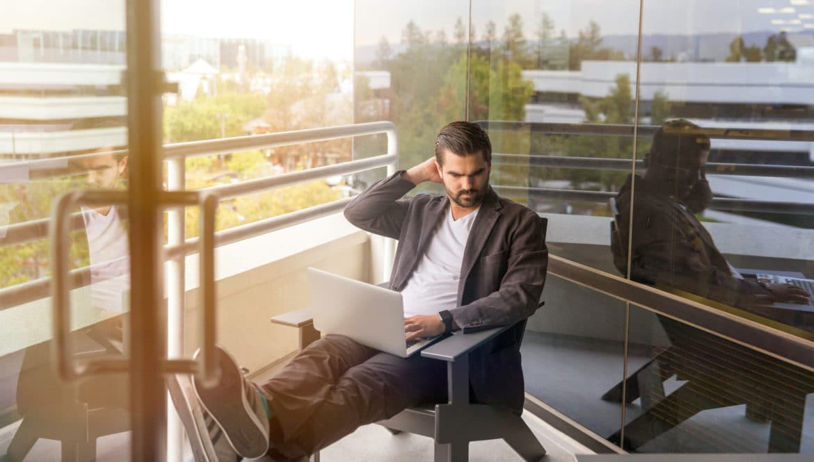 A man using a laptop computer for remote work while sitting on a bench.