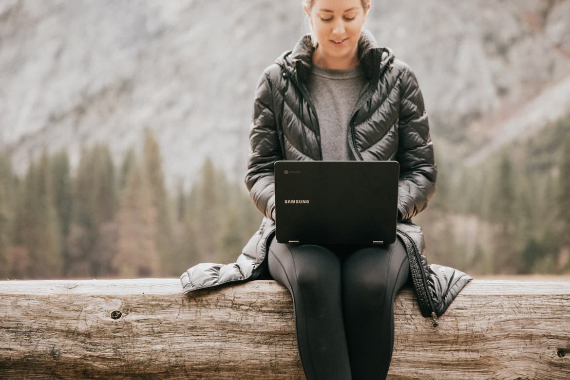 A woman sitting on a log using a laptop, motivated.