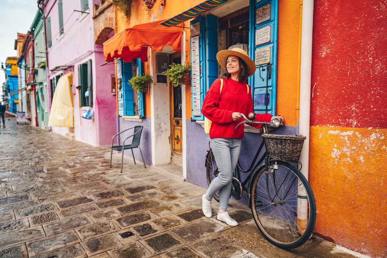 A woman is leaning on a bicycle in front of colorful houses in Burano, Italy during relocation.